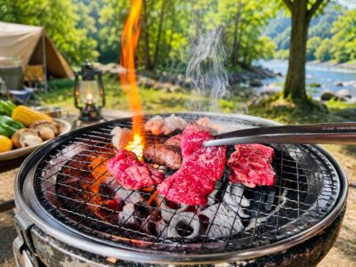 キャンプ場で炭火の上に置かれた網で牛肉を焼いており、炎と煙が立ち上っている様子。