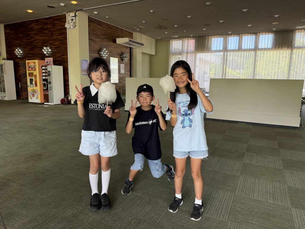 屋内でわたあめを持ち、ピースサインをして並んでいる3人の子供たちの全身写真。